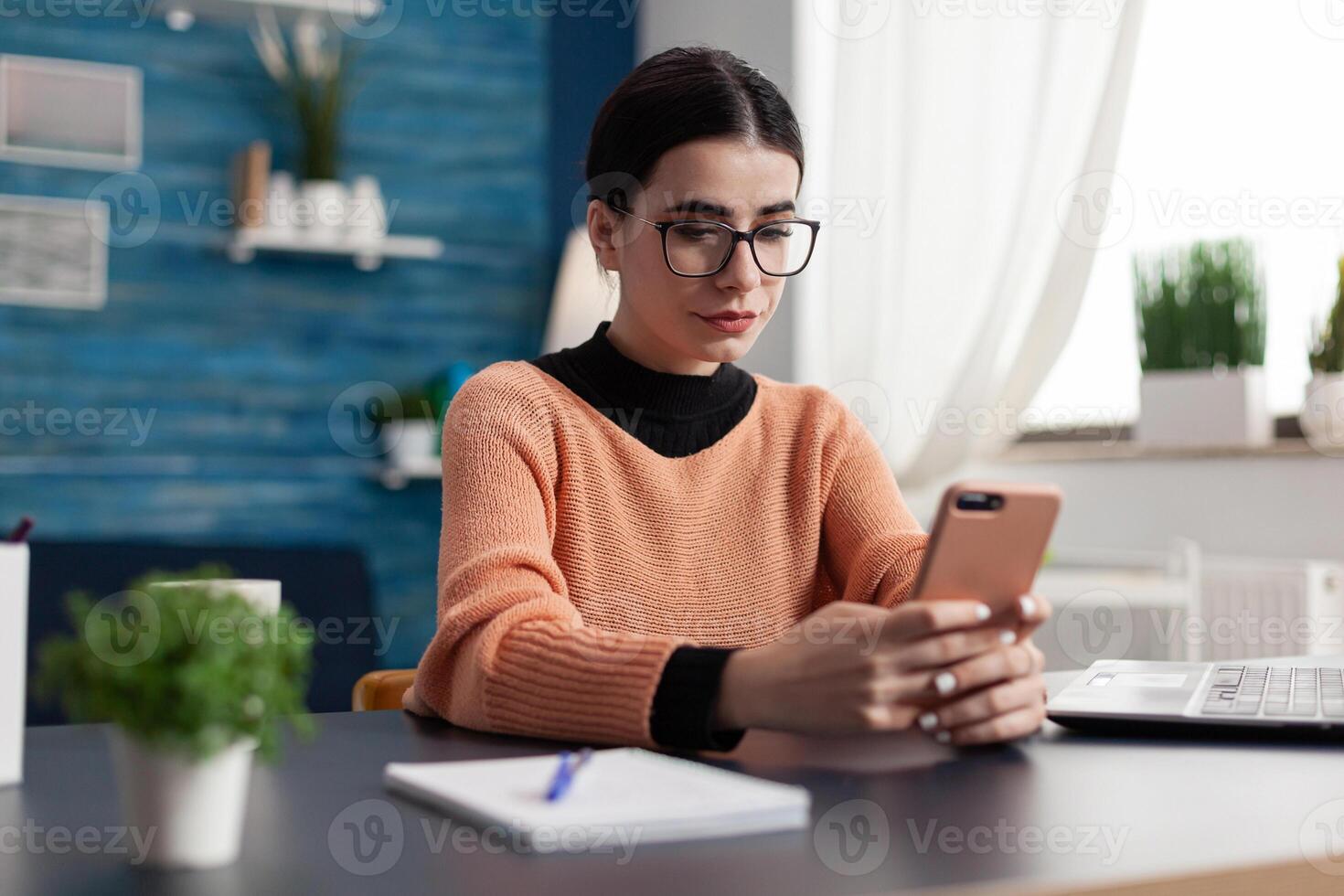 Student looking at smartphone screen while texting message sitting at desk table in living room. Student browsing lifestyle information on internet during coronavirus quarantine photo