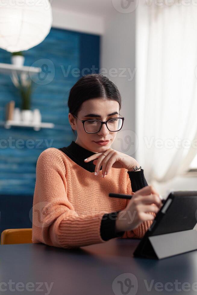 Student artist holding graphic pen in hands drawing illustration sketches on screen of digital tablet computer. Focused woman with glasses sitting at desk table in living room looking at sketch photo