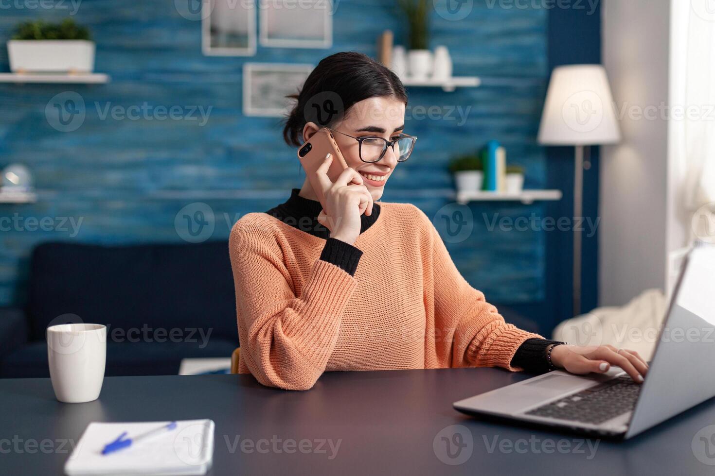 Teenager studying at communication project for university seminar using laptop computer. Student talking on phone with collegue while sitting at desk table in living room photo