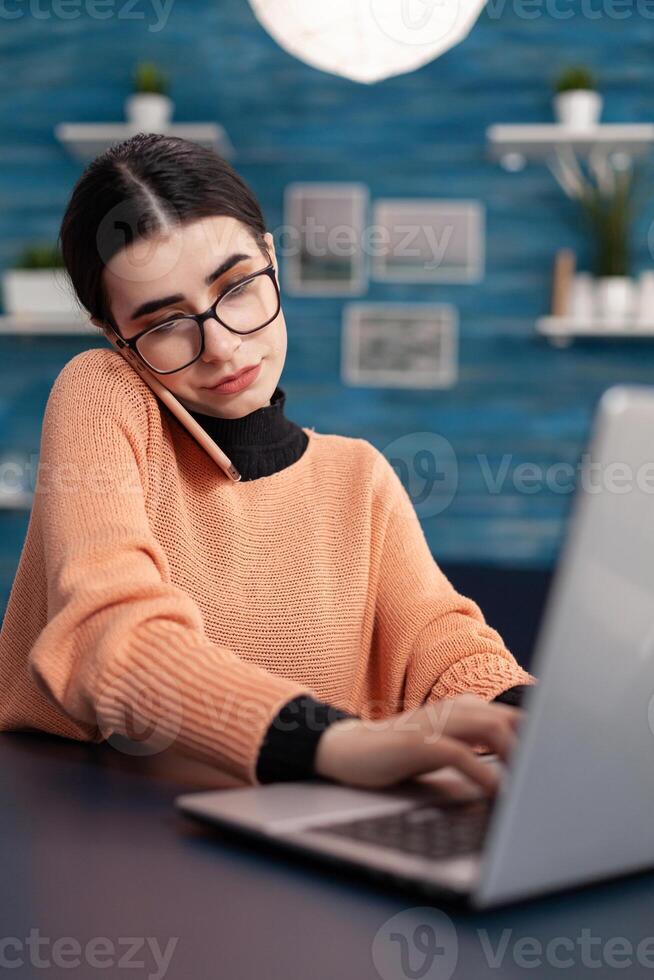 Student browsing information on laptop computer for digital seminar. Teenager woman speaking at phone whit collegue about online university meeting while sitting at desk in living room photo