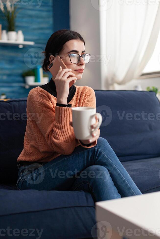 Student talking with her friends about social media using modern smartphone, relaxing alone on couch during coronavirus quarantine. Teenager gossiping while drinking coffee photo