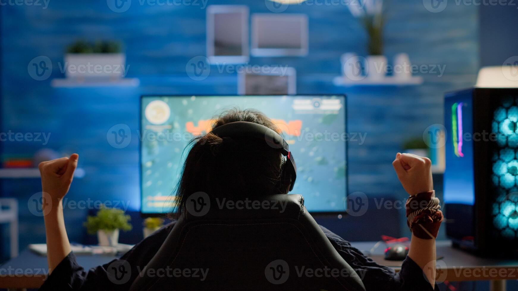 Back view of happy woman gamer winning space shooter game in gaming home room. Cyber performing on RGB powerful computer streaming games using professional headphones for online championship photo