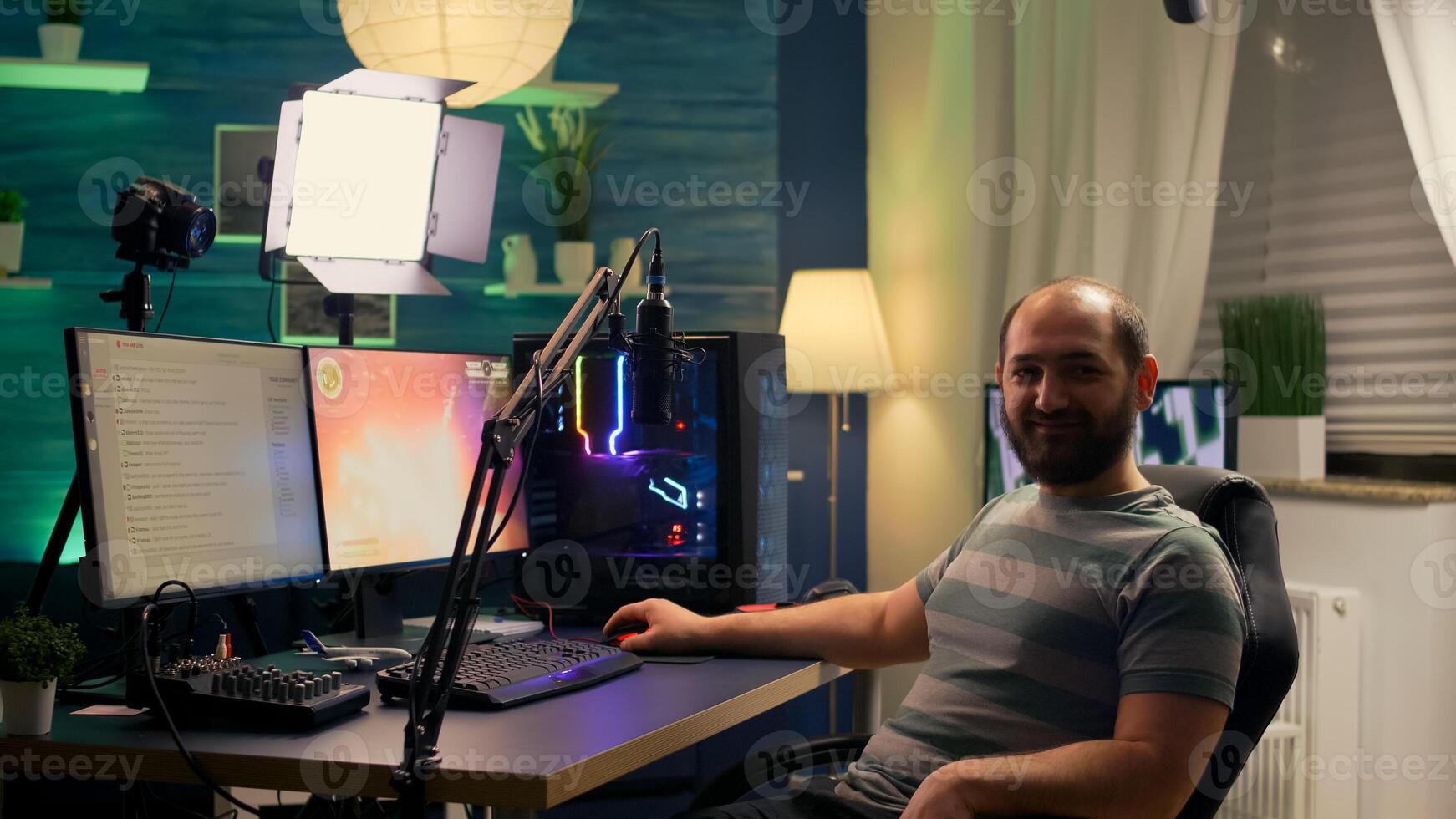 Streamer man looking at camera and smiling while streaming chat is open. Videogamer streamer playing space shooter game at gaming home studio equipped with professional equipment photo