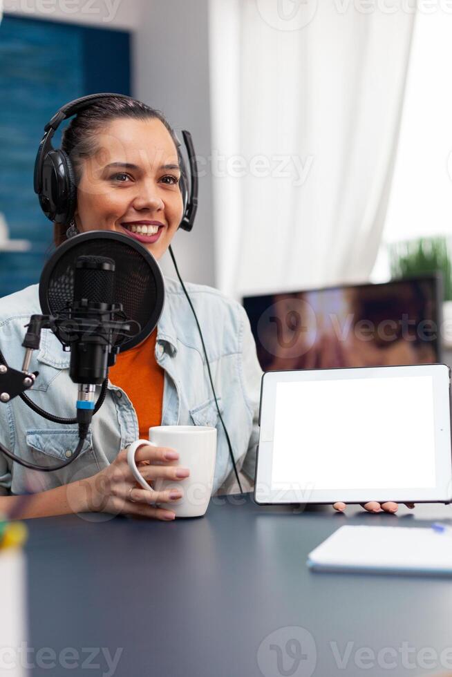 Woman smiling showing digital tablet with white screen. Blogger holding device with mockup template and isolated background while using microphone and headphones to stream podcast. photo