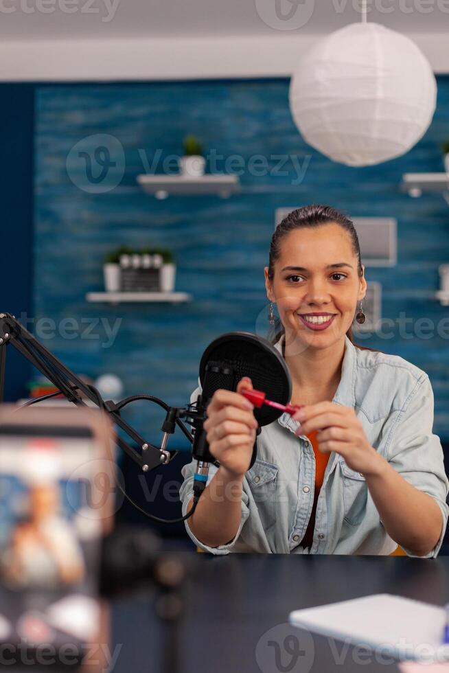 Smiling influencer filming lipstick review with mobile phone. Woman vlogger recording make-up recommendation with smartphone for social media channel. Online broadcasting in home studio. photo