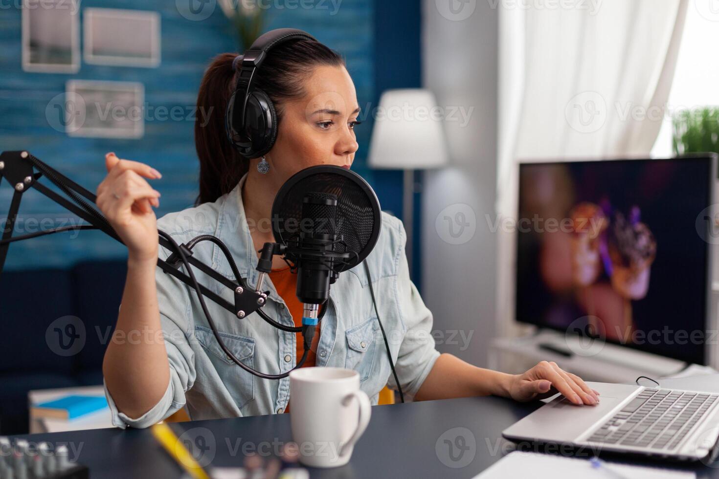 Woman talking on microphone while using laptop. Content creator wearing headphones recording podcast with audio equipment. Social media influencer in home office broadcasting live. photo