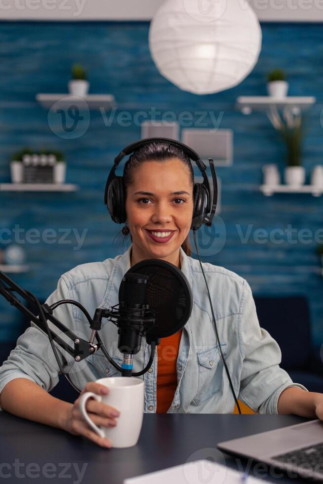 Portait of woman vlogger holding cup and streaming podcast using laptop. Social media influencer recording livestream with headphones and microphone in home office. Smiling blogger podcasting. photo