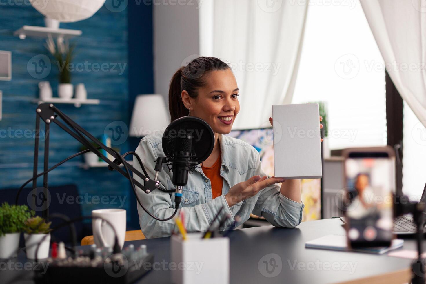 Smiling vlogger presenting white product box in front of camera. Woman content creator recording online vlog review for social media channel. Home recording studio with microphone and laptop. photo