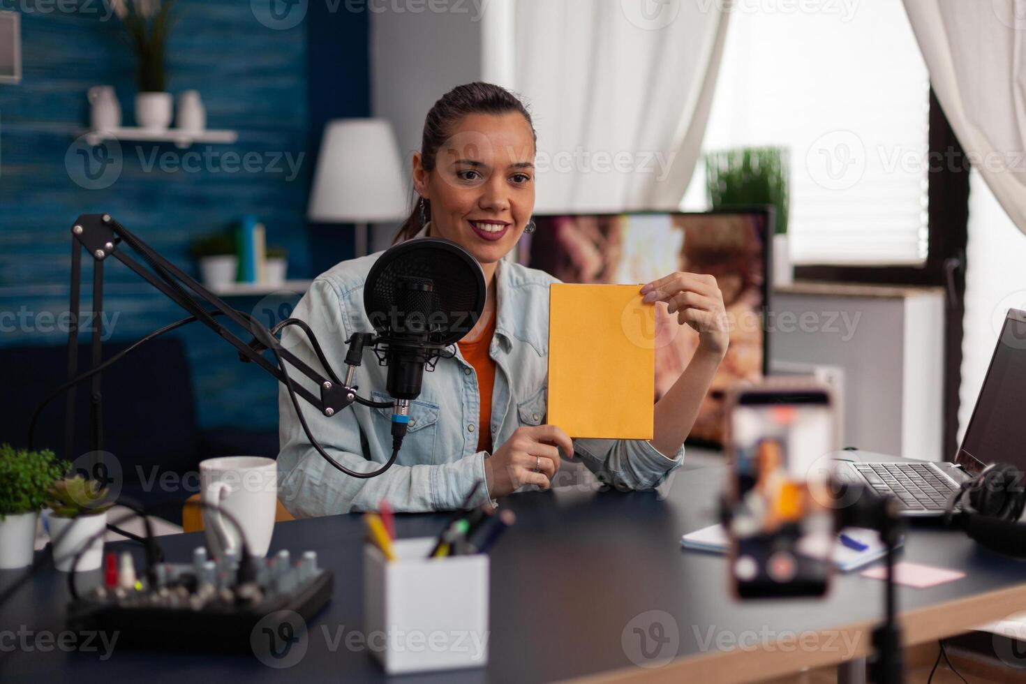 Woman influencer presenting yellow book in front of recording smartphone. Similing content creator broadcasting review for social media channel in home studio with microphone and laptop. photo