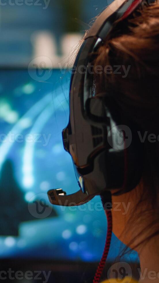 Over the shoulder pro gamer woman playing computer FPS shooter game on championship, talking into headset . Cyber performing on powerful computer in stylish room during gaming tournament photo