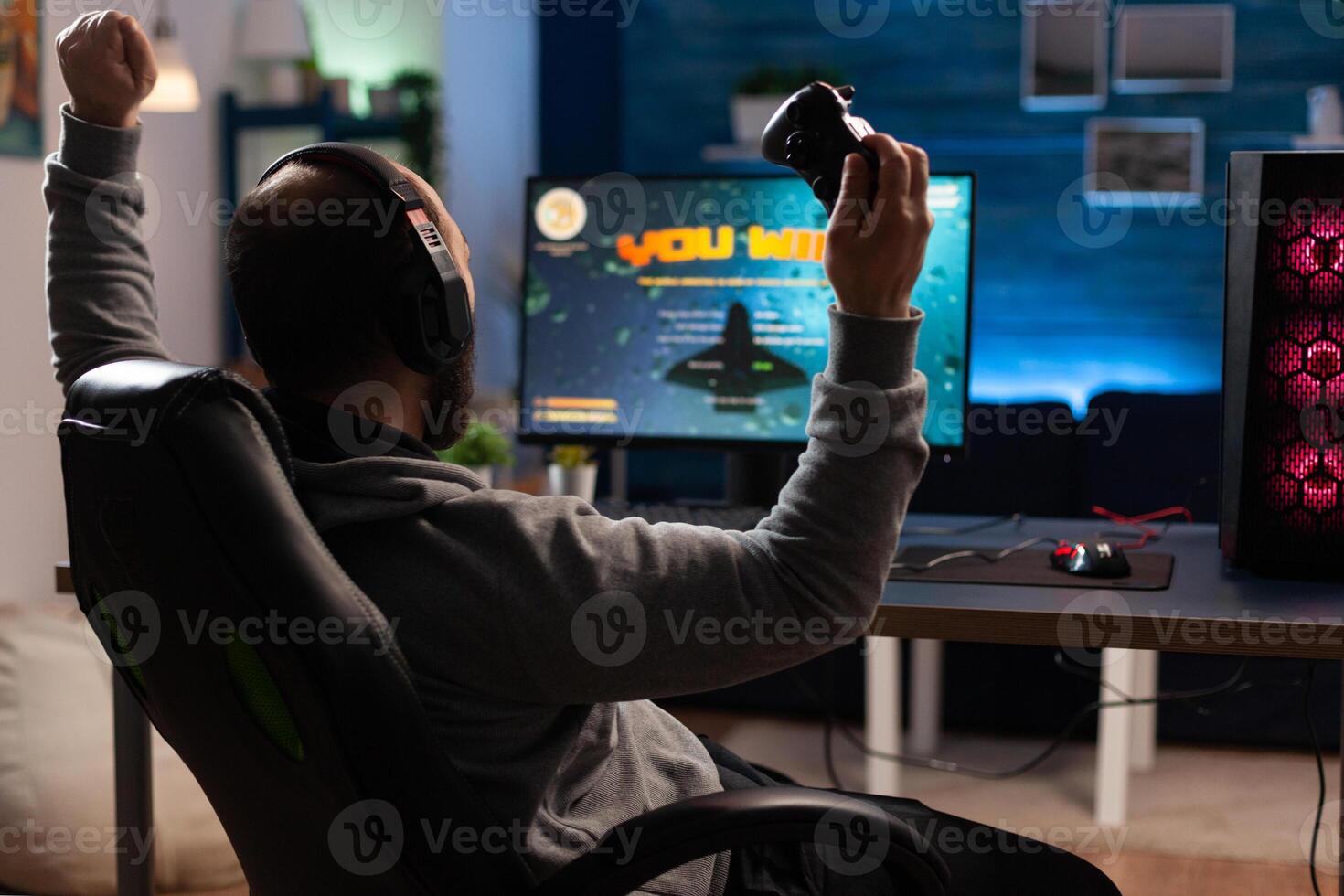 Man winning games with joystick and headphones in front of monitor. Gamer using controller and headset, playing online games on computer. Person celebrating game win for leisure. photo