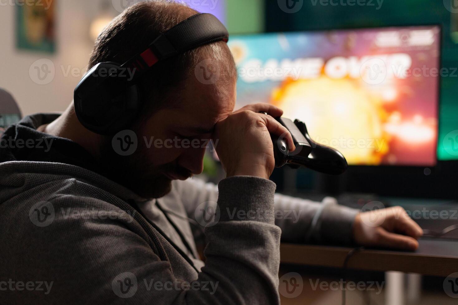 Sad gamer losing games after playing with controller and headphones on computer. Man feeling disappointed about lost game, using controller and headset. Player gaming with equipment photo