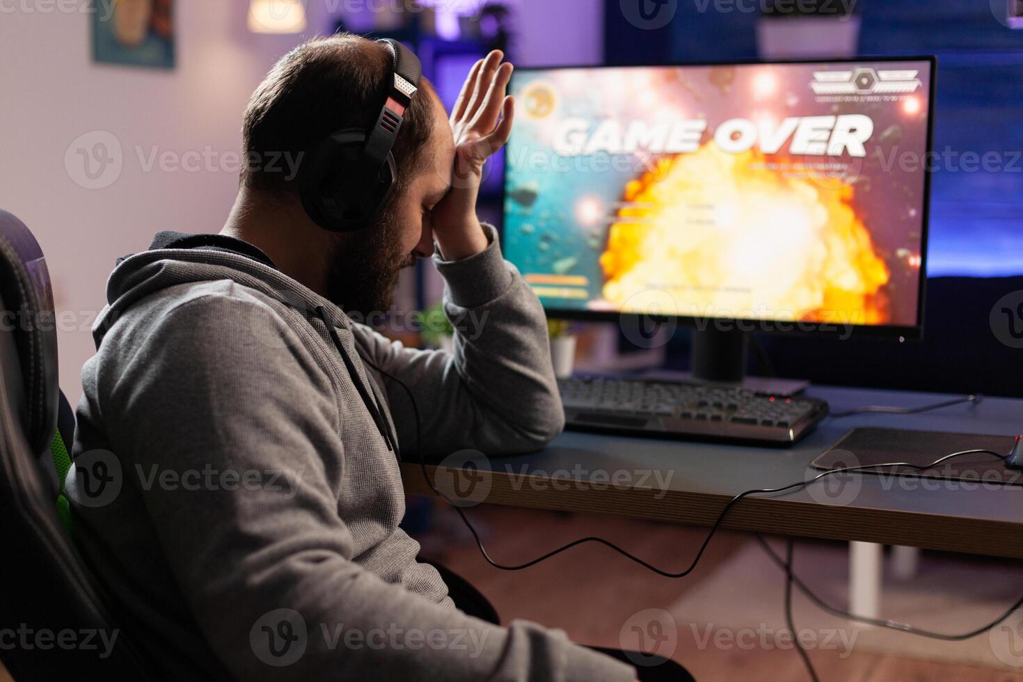 Sad gamer losing games play on computer. Man playing online games with headphones and controller, feeling disappointed about lost game. Player using joystick and monitor. photo