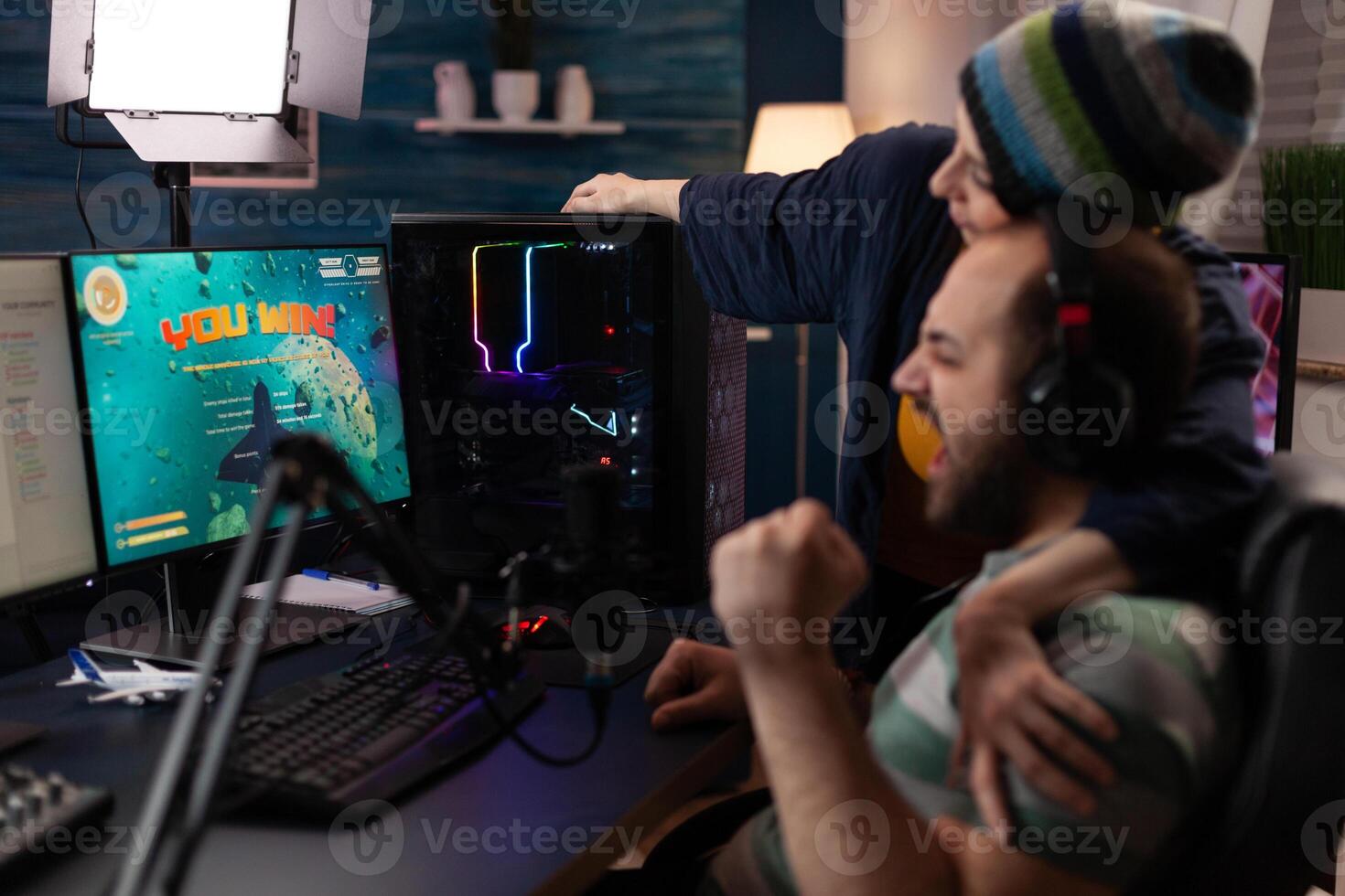 Couple celebrating games win on live stream. Woman feeling happy about man winning gameplay while he is streaming online with chat on computer. Streamer using gaming equipment photo