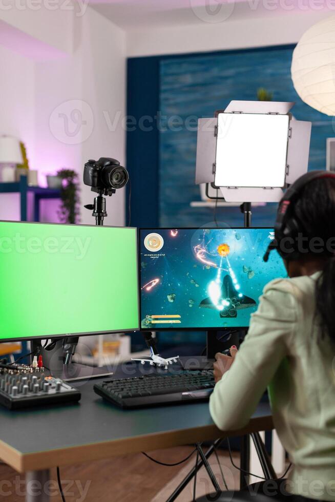 Gamer woman playing online videogames during gaming championship loooking at mok up green screen chroma key computer with isolated display. Young player streaming space shooter game using joystick photo