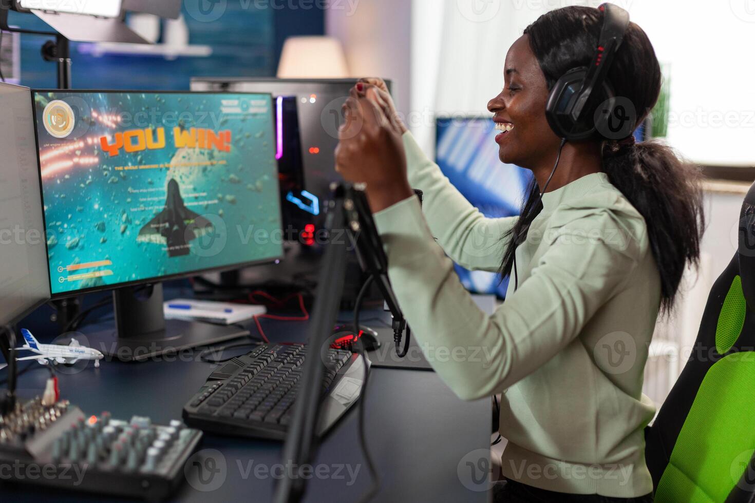 Happy african american woman gamer wearing headset winning videogames competition talking into microphone with players. Excited plater playing space shooter games using RGB computer. Live streaming photo