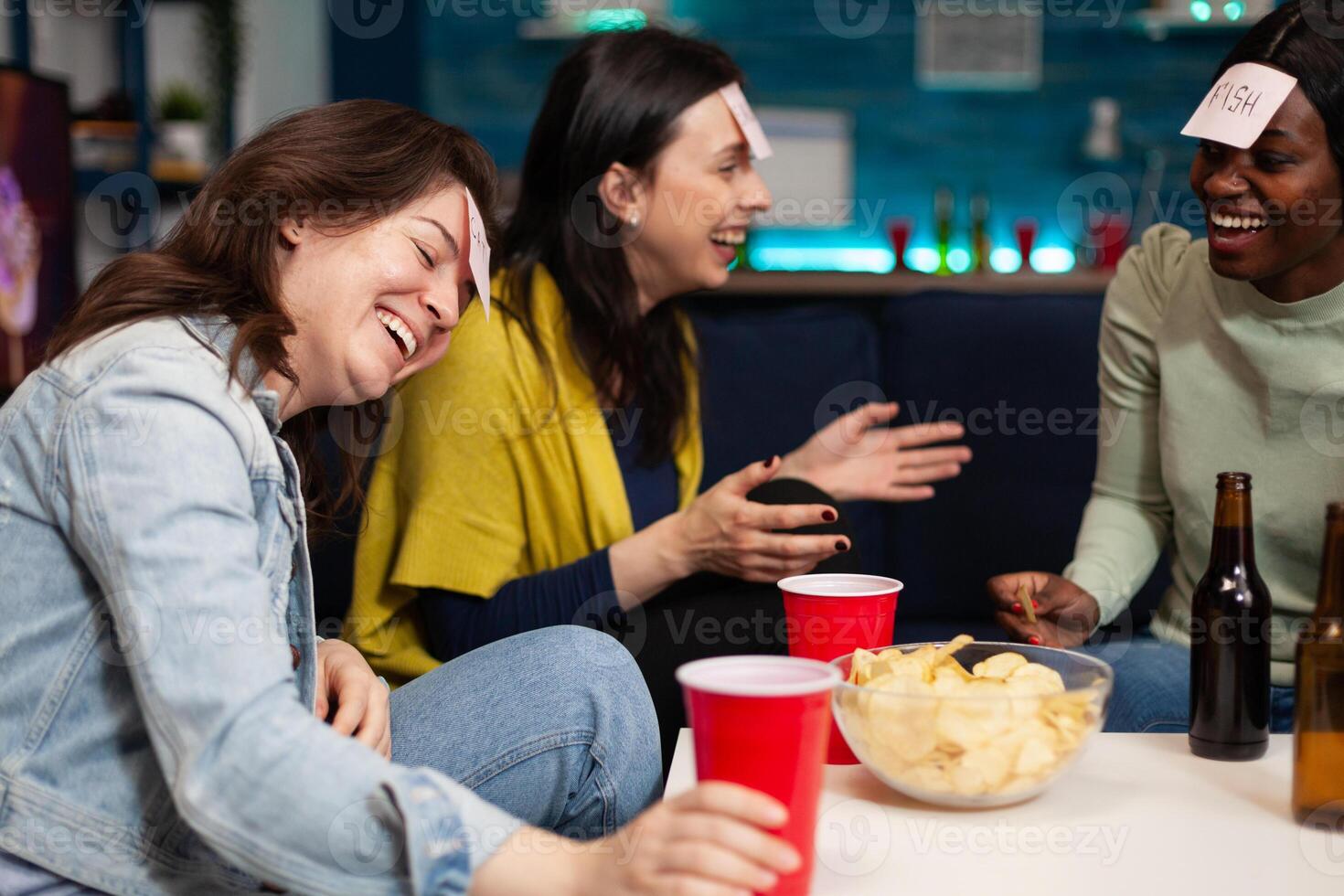 Laughing multi-ethnic friends playing who i am game using stickey notes enjoying spending time together during wekeend party at home. Group of people drinking beer guessing correct answer photo