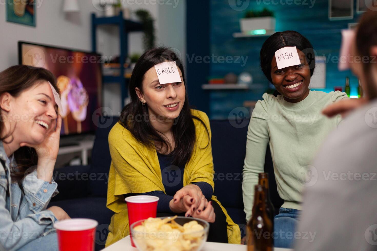 Multi-ethnic friends with stickey notes on forehead playing guess who game enjoying spending time together hanging out at home in living room. Group of people having fun during evening party photo