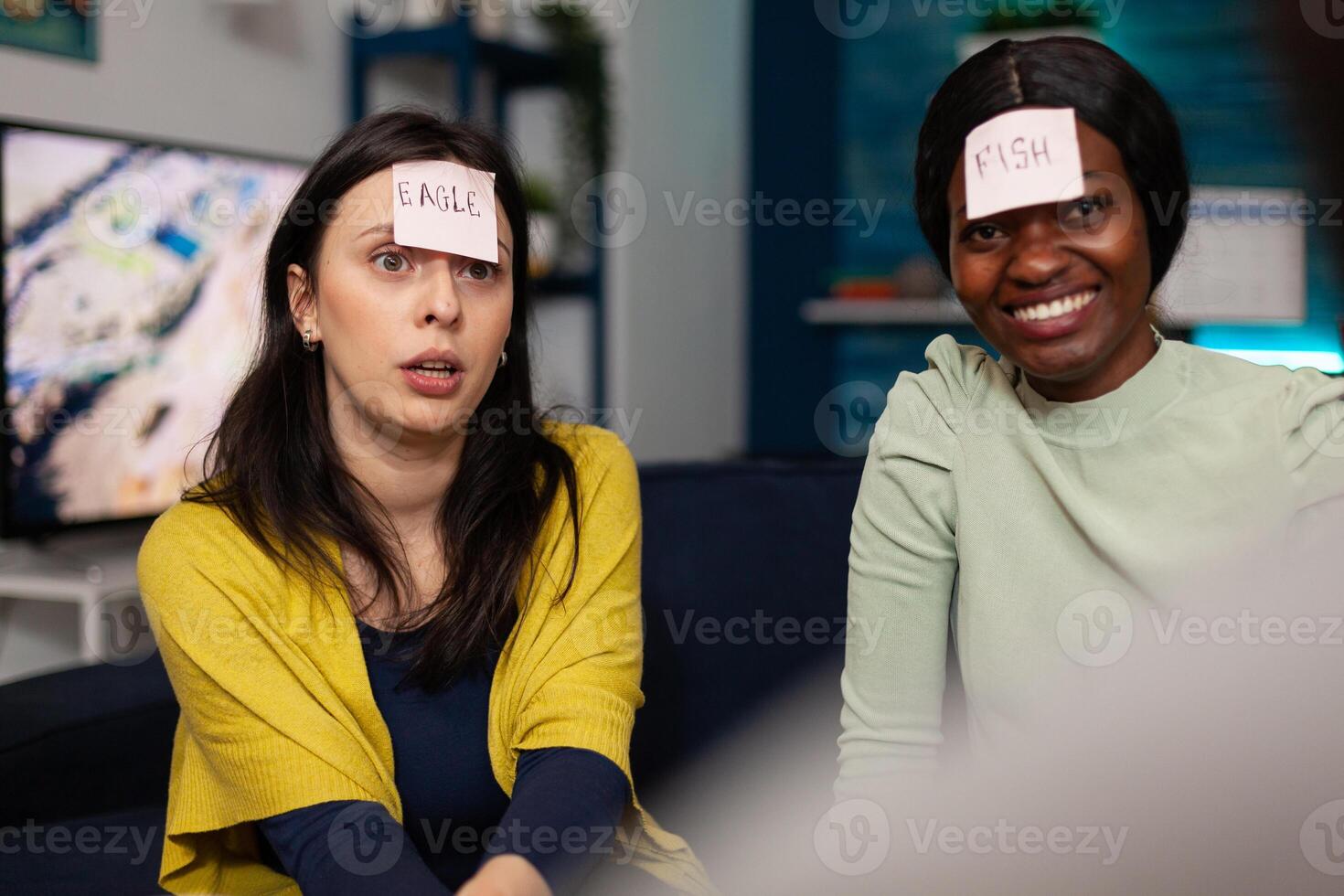 Portrait of multiethnic friends with stickey notes on forehead playing who i am game enjoying spending time together hanging out at home in living room. Group of people having fun during wekeend party photo