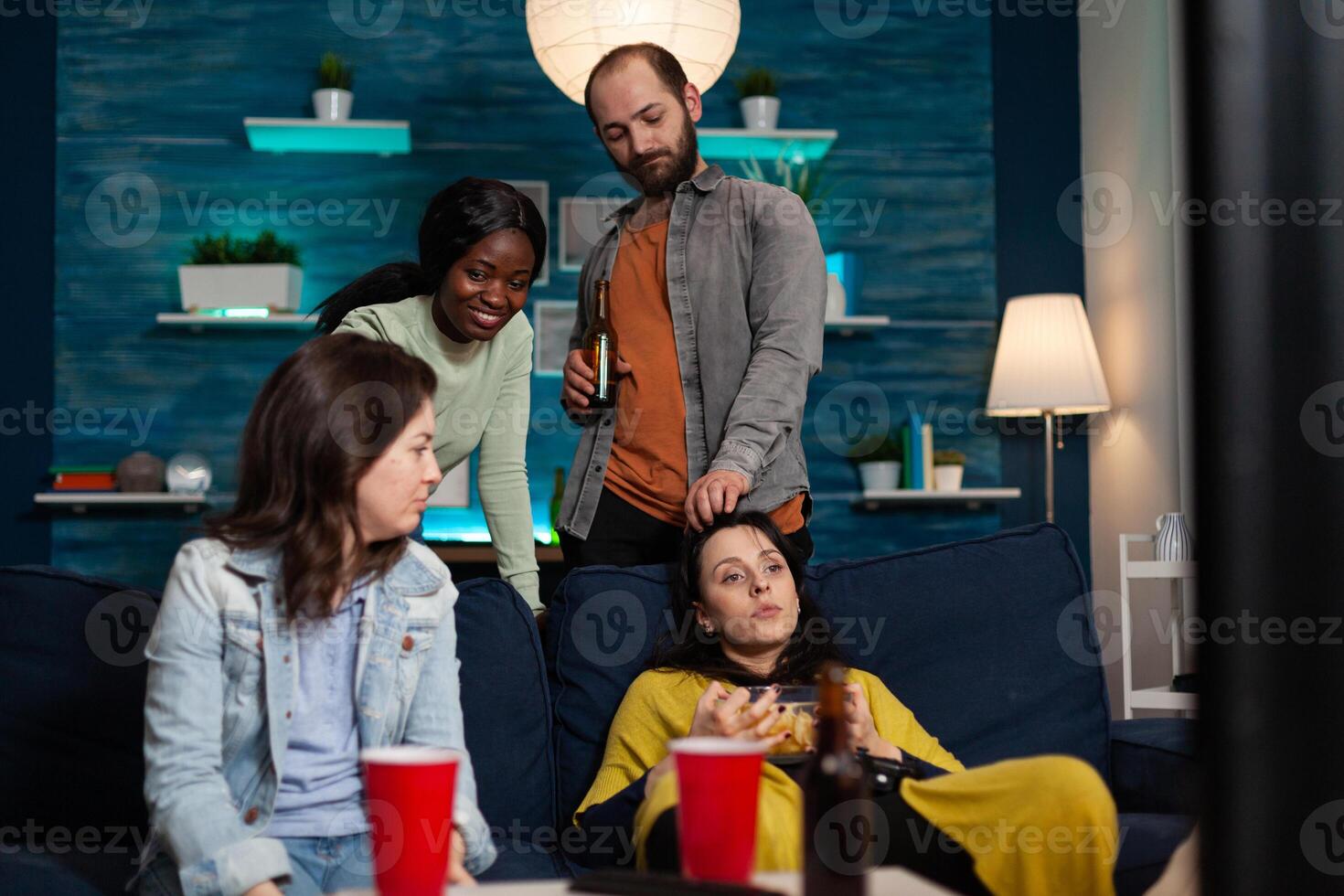 Multi-ethnic friends sitting on sofa looking at television watching online chill movie enjoying wekeend night at home. Group of people eating snack, drinking beer having fun. Hanging out concept photo