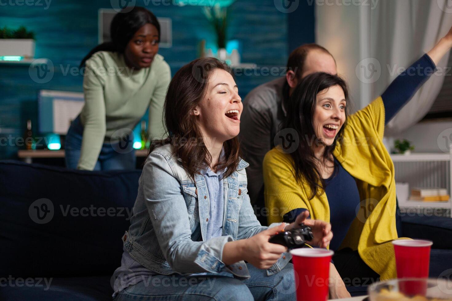 Happy excited women playing videogames using gaming controller winning online competition enjoying spending time in living room. Multi-ethnic friends having fun together during wekeend party photo