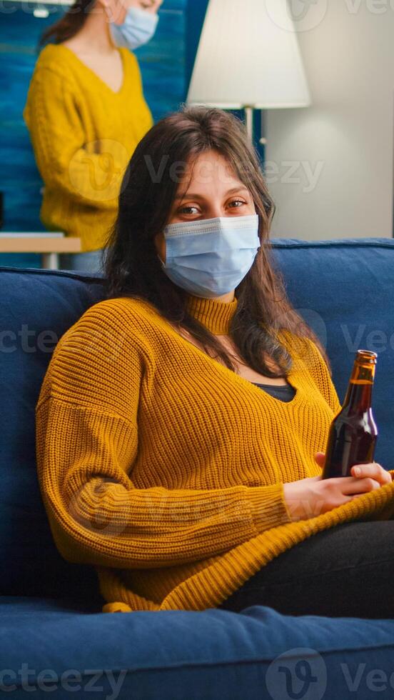 Woman relaxing with friends sitting on couch wearing face mask against spreading coronavirus during global pandemic holding beer bottle looking at camera. People socializing during covid 19 outbreak photo