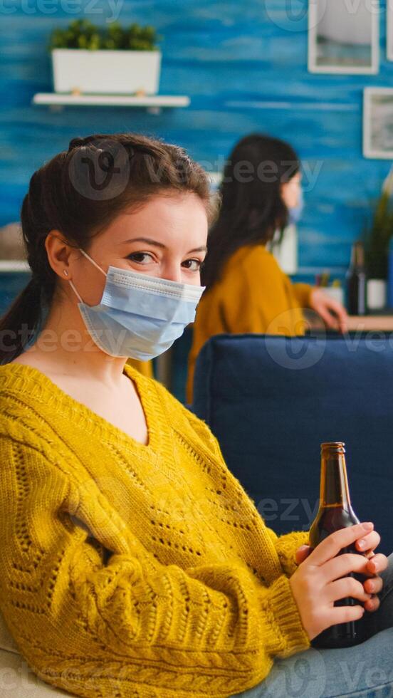 Pretty lady taking off protection face mask looking at camera drinking beer spending good time with friends in living room respecting social distance. People socializing during covid 19 outbreak photo