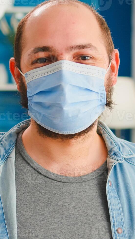 Portrait of man putting on protection mask looking at camera spending good time with friends in living room respecting social distance in global pandemic. People socializing during covid 19 outbreak photo