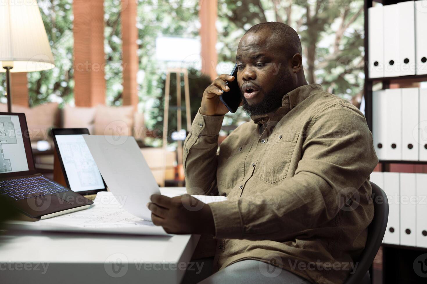 Professional businessman collaborating with contractors on phone call to plan office spaces, using blueprints and digital tools for a modern layout. Communicating interior ideas. photo