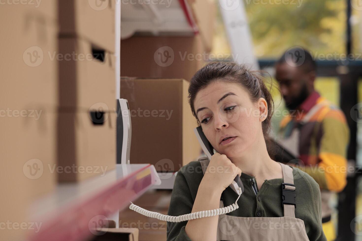 Storage room employee talking at landline phone with remote business owner before start working at clients packages shipment. Worker doing products quality control in warehouse. Delivery concept photo