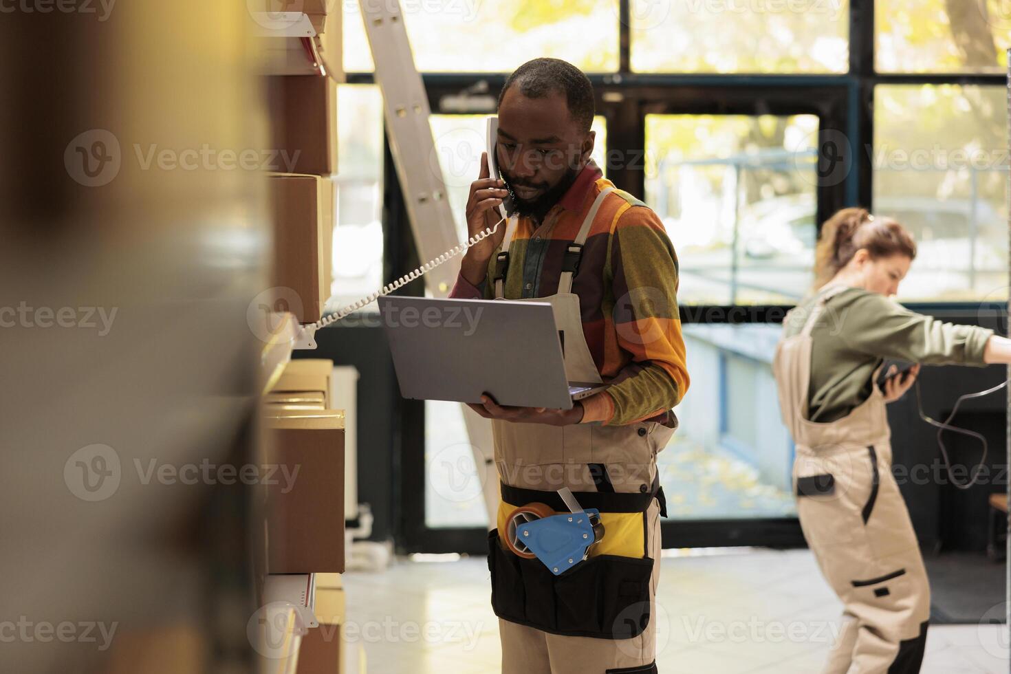 Warehouse worker talking at landline phone with remote manager, looking at customers order on laptop computer. African american supervisor preparing packages for delivery in storage room photo