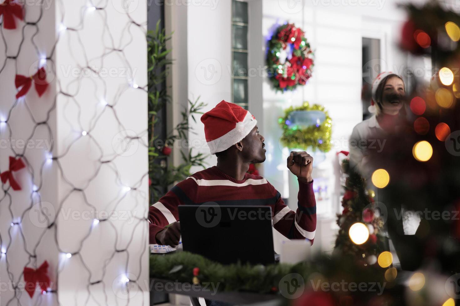 Worker tasked with stressful job duties bothered by background noise made by coworkers laughing while enjoying Christmas holiday season. Employee working on laptop, surrounded by in xmas ornaments photo