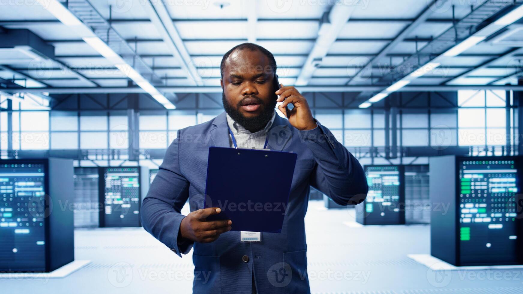 IT professional looking over data center checkup checklist paperwork, talking on phone in server farm. African american man surrounded by server rigs, chatting during telephone call, camera B photo
