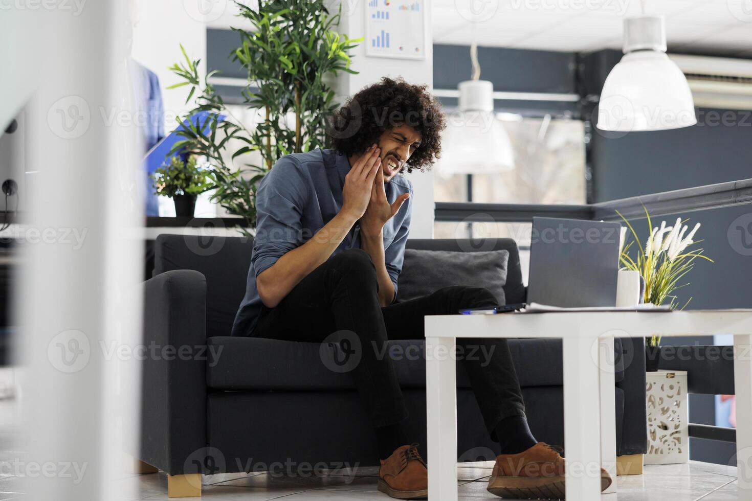 Young arab business manager suffering from toothache in start up office. Company employee having tooth pain while working at strategy on laptop in corporate coworking space photo