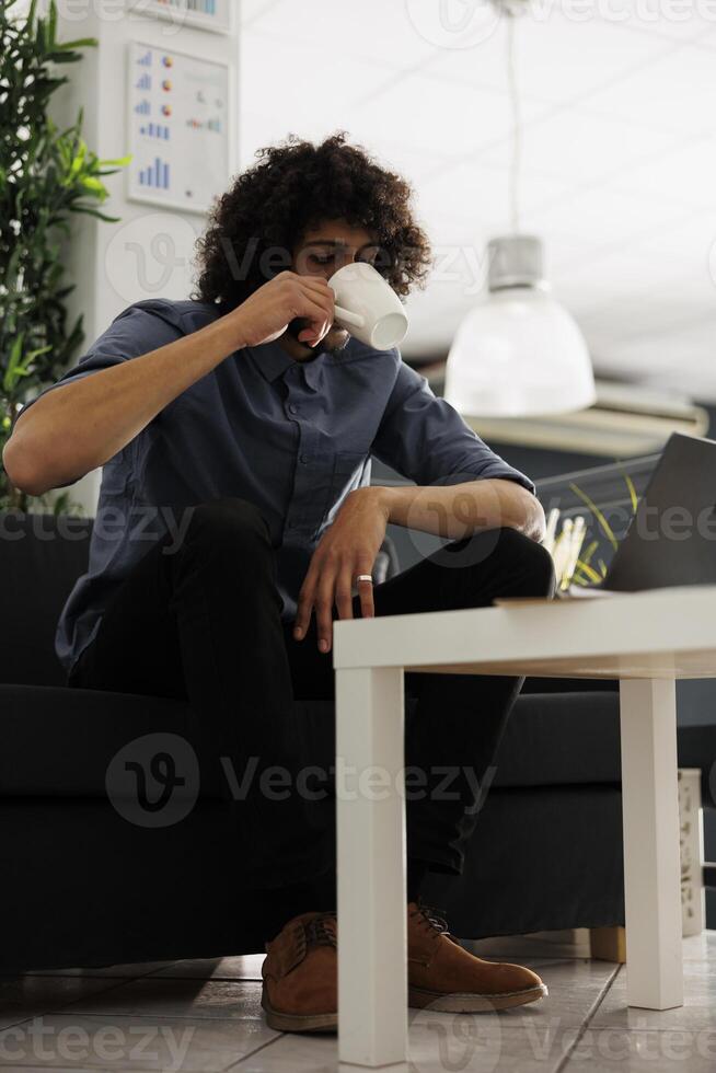 Young arab entrepreneur drinking coffee while sitting on couch in modern business office. Company executive manager enjoying tea while working on laptop in coworking space photo