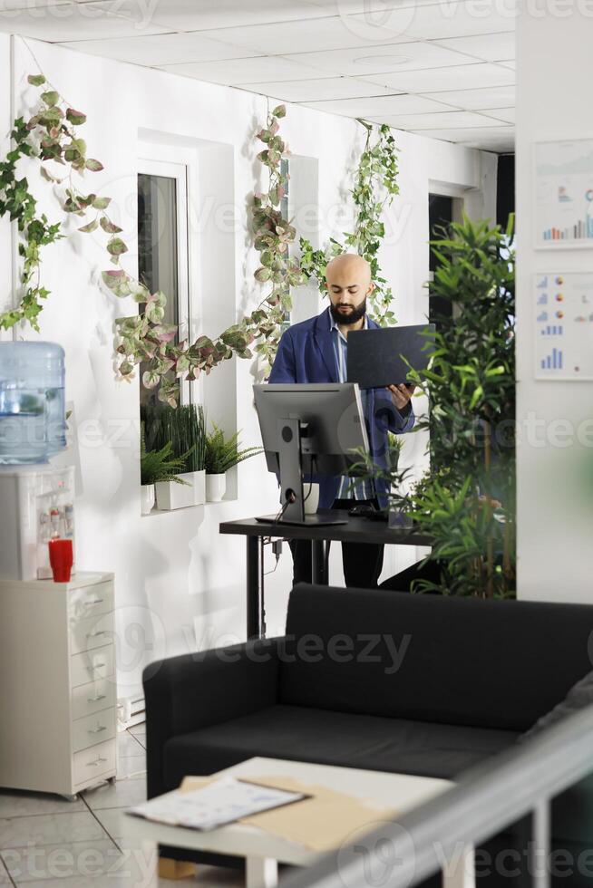 Arab executive manager holding laptop and comparing data on computer while working in start up office. Unicorn business employee standing near workplace desk and analyzing report photo