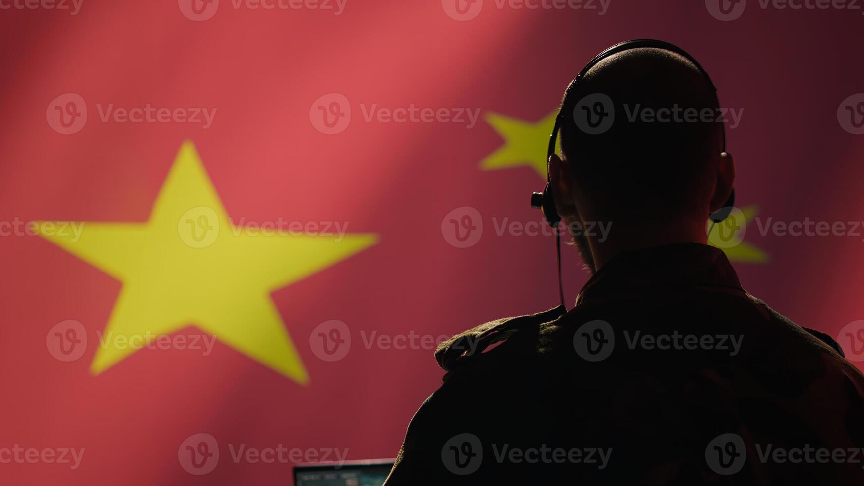 Chinese army military dispatcher looking at surveillance transmission on laptop, relaying informations for troops using headset, isolated over fluttering China flag background, camera B photo