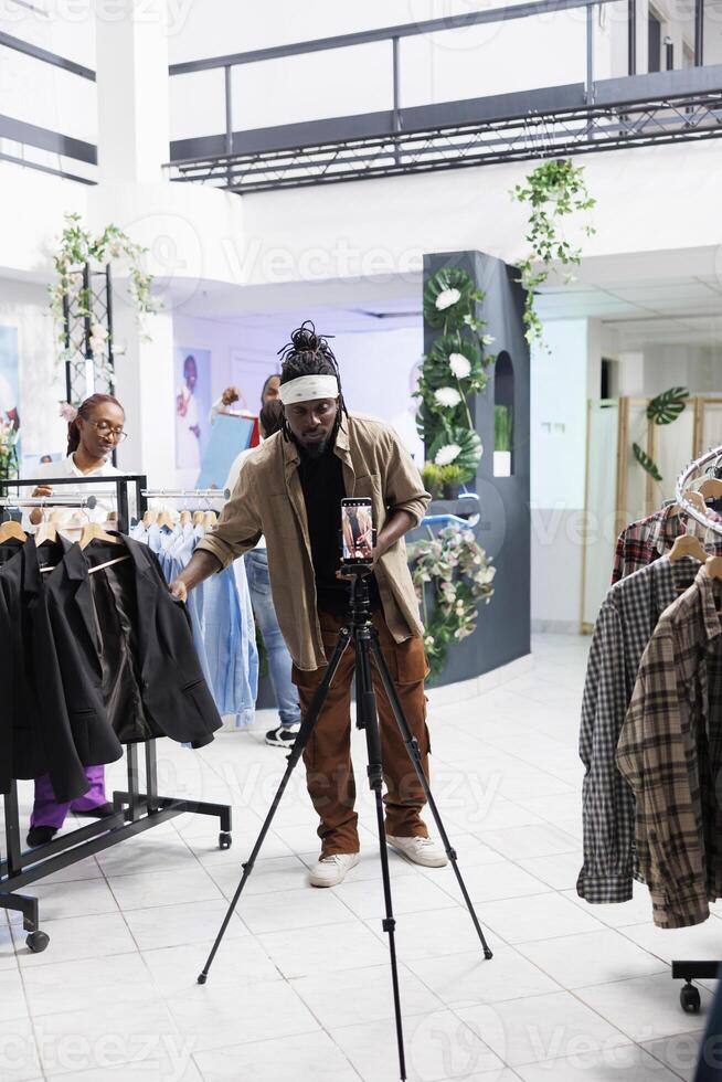 African american social media influencer promoting formal wear collection on smartphone camera in clothing store. Blogger showing jackets while filming live for followers in boutique photo