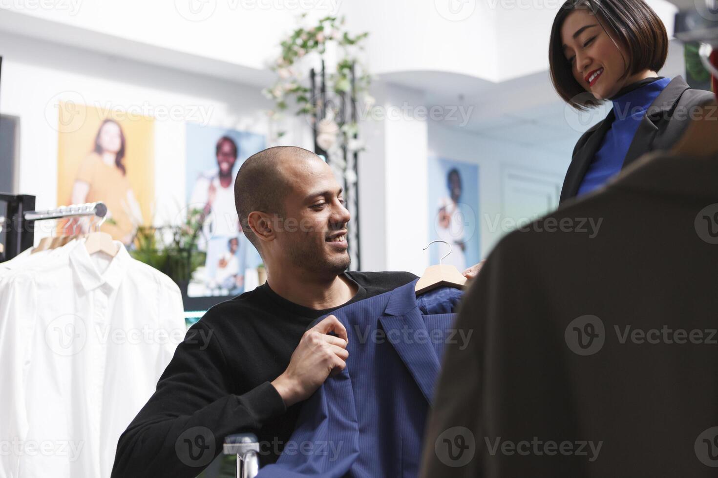 Inclusive boutique asian woman worker offering help to arab customer with disability shopping for formal garment. Buyer in wheelchair holding suit on hanger and discussing choice with seller photo