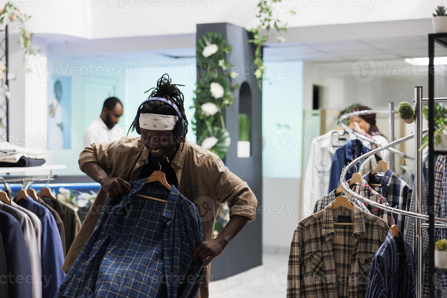 Happy african american man in headphones holding shirt to body while choosing clothes in store. Cheerful buyer listening to music in earphones while shopping for outfit in mall photo