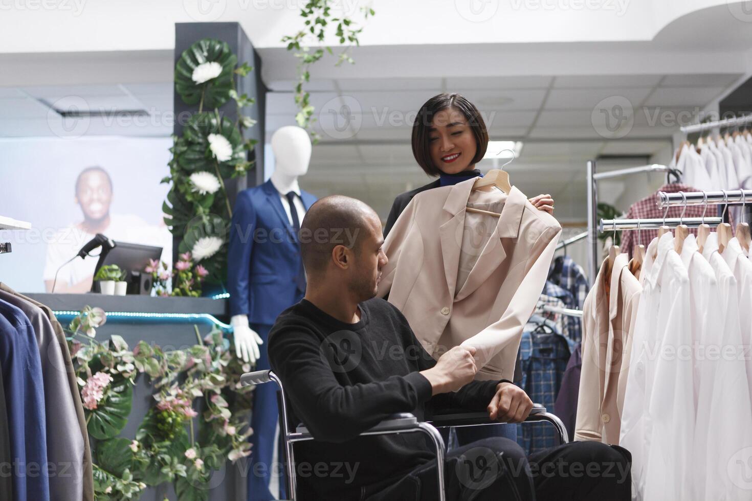 Inclusive clothing store asian woman employee consulting arab man client in wheelchair. Retail center boutique seller assisting buyer with physical impairment in checking jacket photo