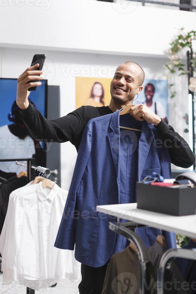 Clothing store smiling customer showing jacket on hanger while chatting in videocall using smartphone. Fashion blogger showing apparel while streaming online with mobile phone photo