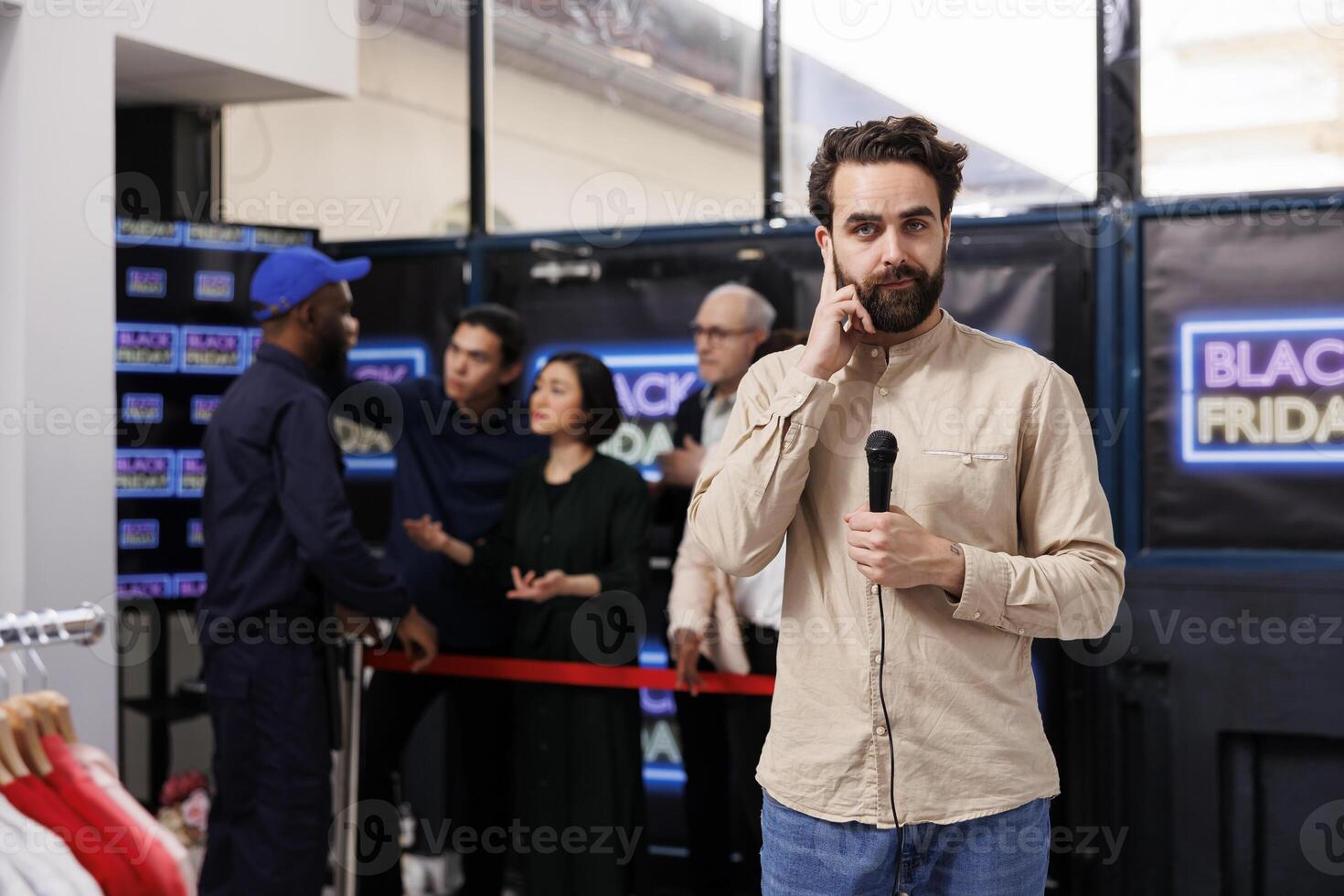 Young man TV reporter checking earphone looking at camera while making Black Friday news episode. Broadcast journalist reporting about crowd of shoppers waiting to get into store during seasonal sales photo