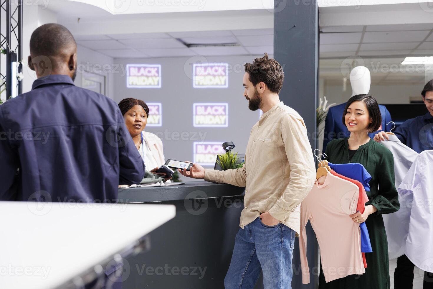 Man standing in line making contactless smartphone payment, paying for purchases with electronic credit card while shopping on Black Friday. Diverse people holding clothes waiting at cash regiter desk photo