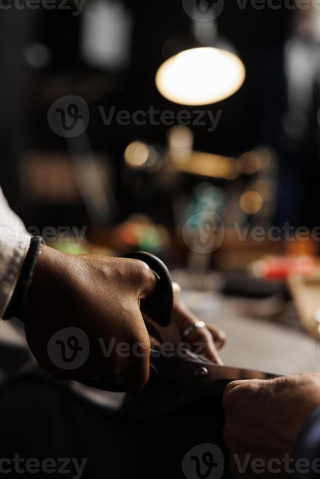 Extreme close up of professional dressmaker using scissors to cut textile fabric, helped by friendly coworker. Experienced couturiers in premium atelier shop studio working on customer comission photo