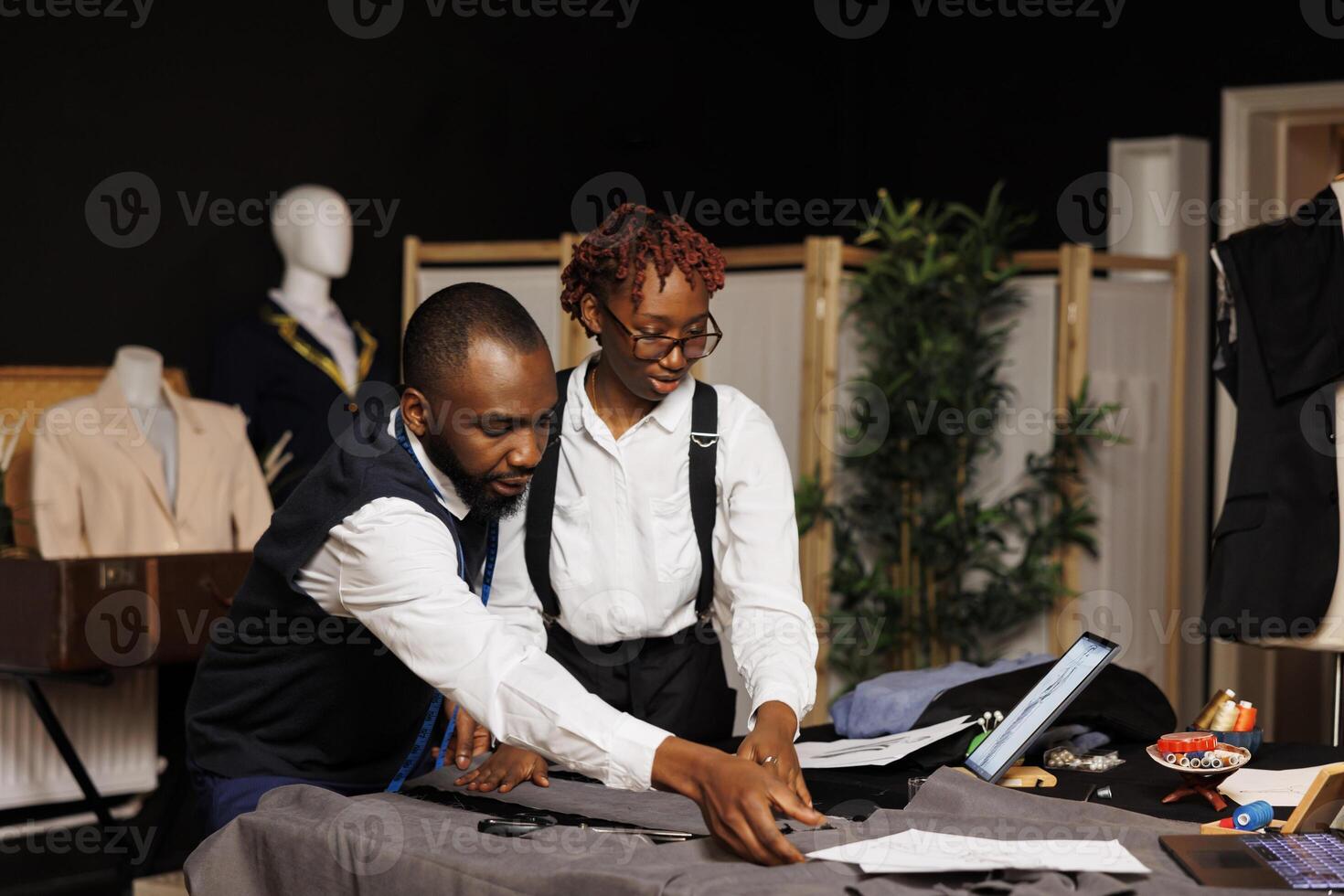Refined masterful tailor team cutting fabric for bespoke modern fashion collection in dark studio. Elegant dressmakers manufacturing attire following dresses sketches photo