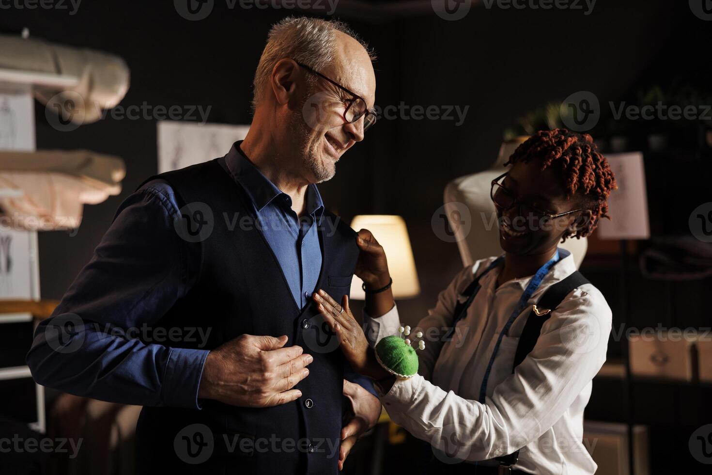 Adept professional couturier helping happy client with premium quality handmade suit comission. Meticulous seamstress doing additional needlework needed on bespoke suit vest worn by senior man photo