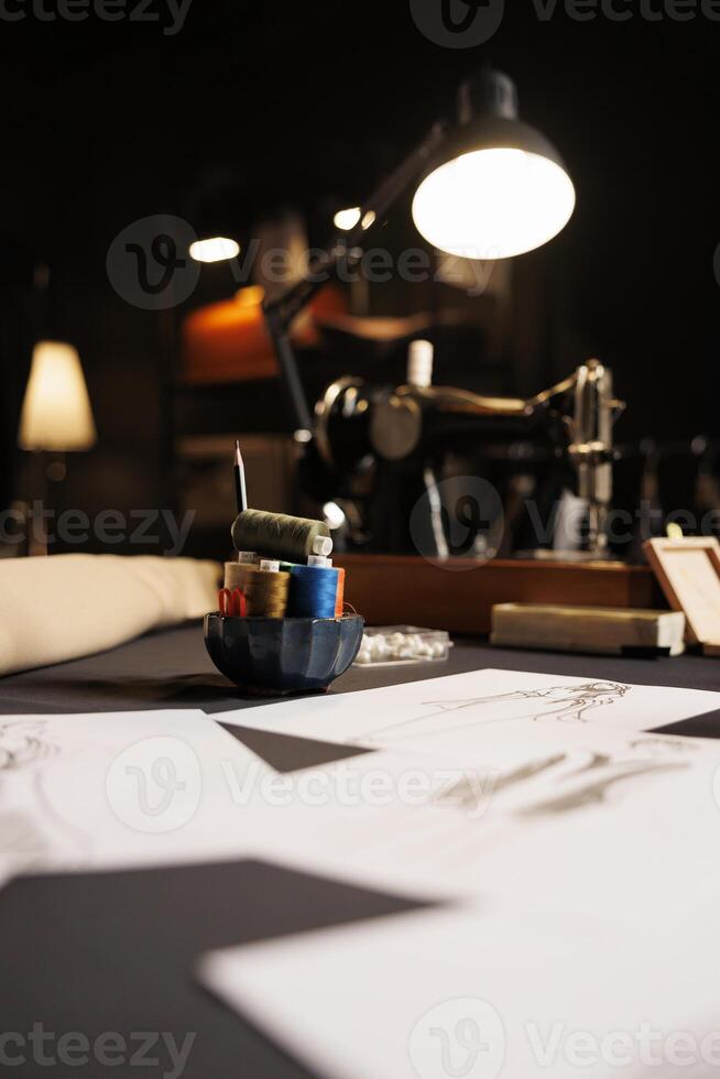 Table full of fashion designs on paper and sewing instruments in tailoring studio. Professional atelier shop workspace with sketch drawings, thread spools, sewing machine photo