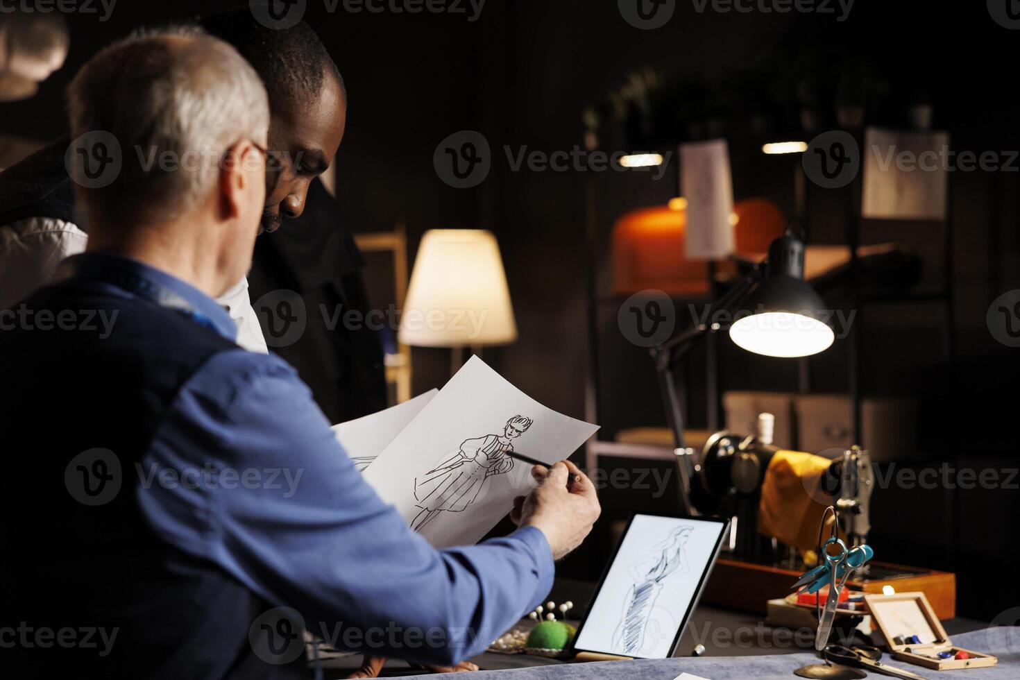 Stylish fashion designers drawing fashionable chic dress sketches, preparing for upcoming fashion design runway show. Bespoke precise tailoring in dark atelier by master and apprentice photo