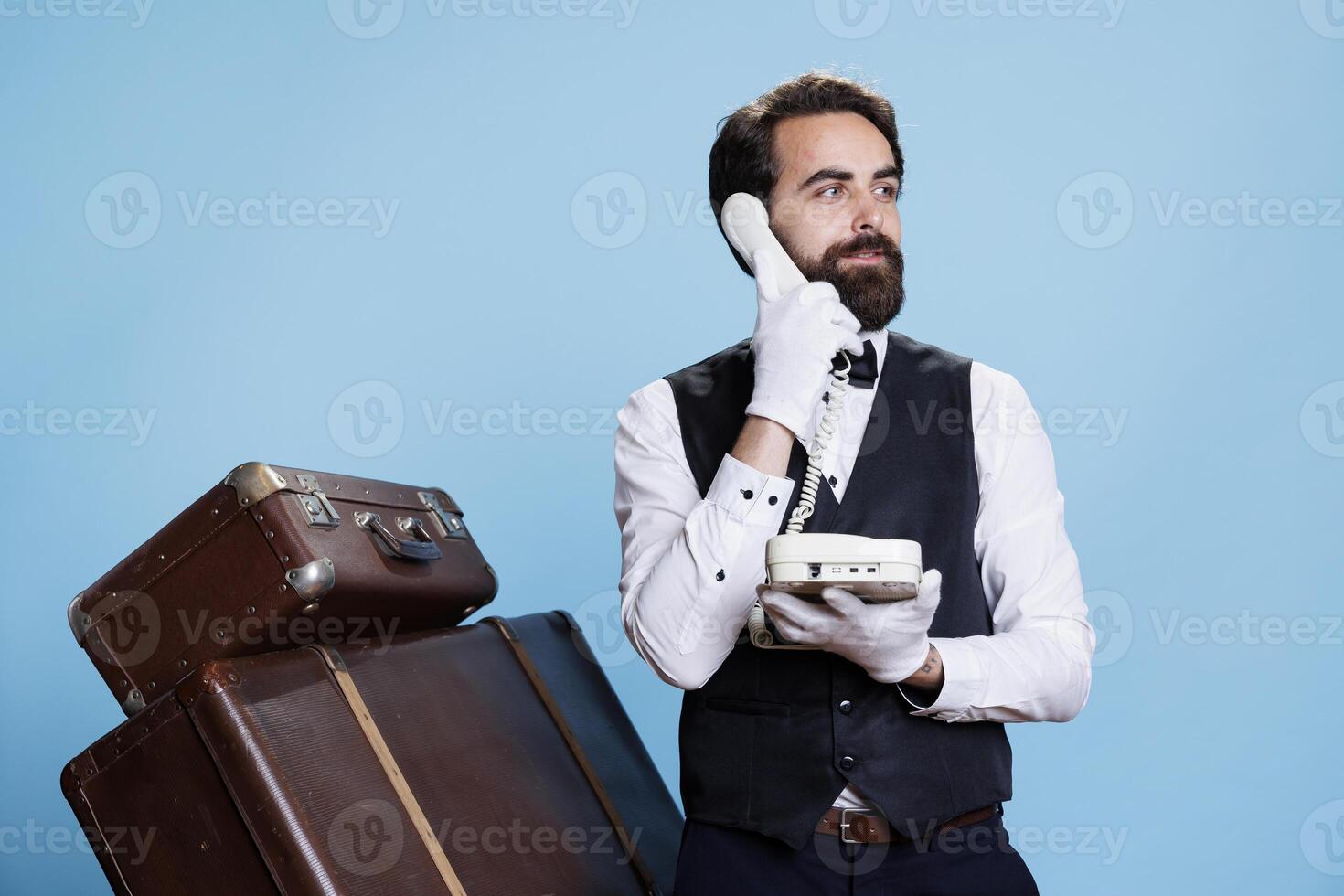 Classy bellhop interacts with tourists about facilities as they make calls on landline phone. Professional competent doorman using wired telephone to take inquiries for aristocratic job. photo